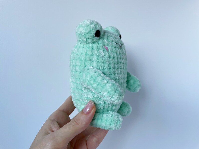 May include: A soft, green crocheted frog plush toy with black eyes and a pink mouth.