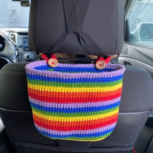 May include: A rainbow striped crocheted hanging basket with wooden button accents. The basket is hanging from the headrest of a car seat.