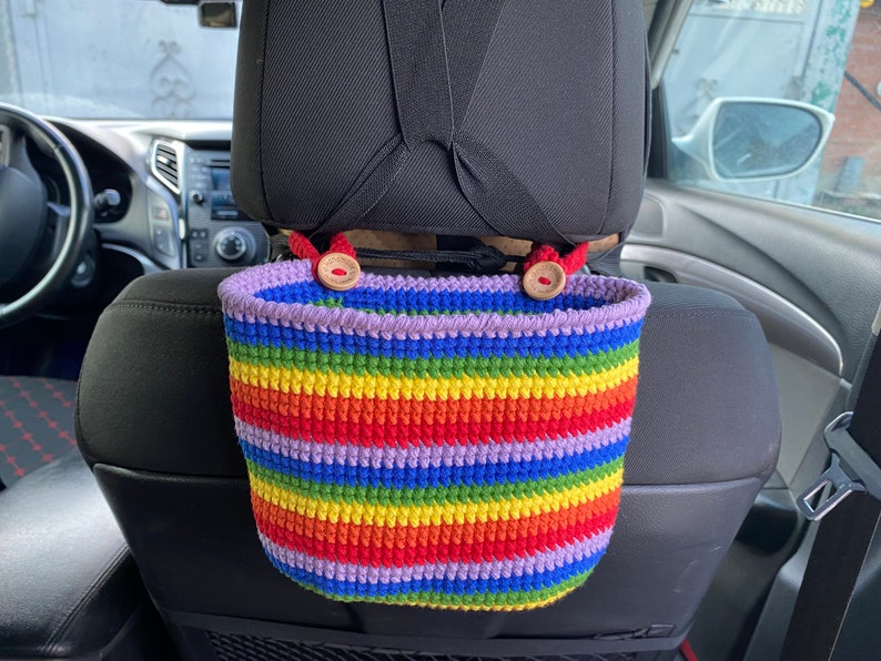 May include: A rainbow striped crocheted hanging basket with wooden button accents. The basket is hanging from the headrest of a car seat.