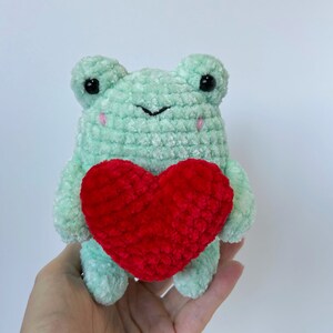 May include: A green crocheted frog holding a red crocheted heart. The frog has black eyes and pink cheeks.
