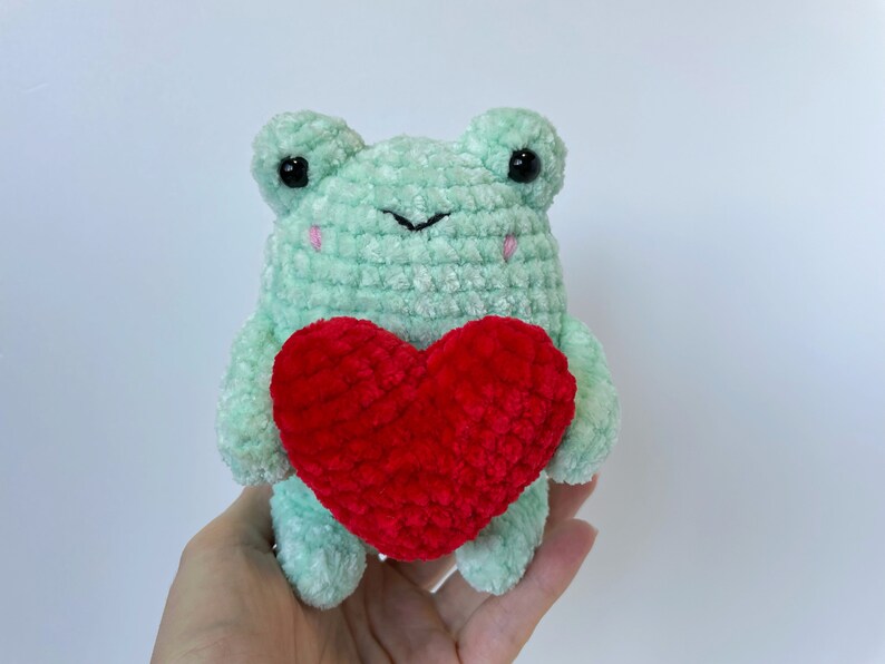 May include: A green crocheted frog holding a red crocheted heart. The frog has black eyes and pink cheeks.