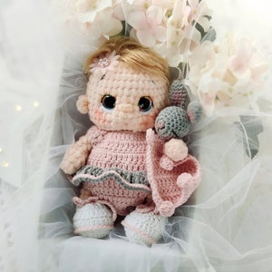 Pu&ograve; includere: Una bambola all'uncinetto con abiti rosa e grigi e un coniglietto di uncinetto grigio tra le braccia. La bambola &egrave; seduta in una scatola bianca con uno sfondo di tulle bianco.