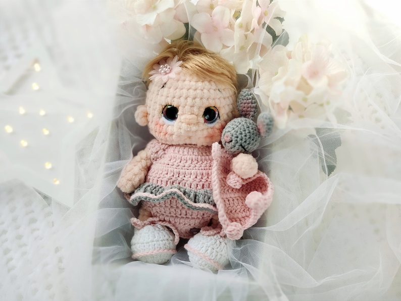 Pu&ograve; includere: Una bambola all'uncinetto con abiti rosa e grigi e un coniglietto di uncinetto grigio tra le braccia. La bambola &egrave; seduta in una scatola bianca con uno sfondo di tulle bianco.