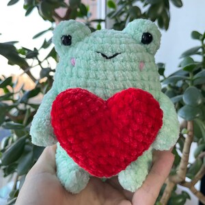 May include: A green crocheted frog holding a red crocheted heart. The frog has black eyes and a black nose.
