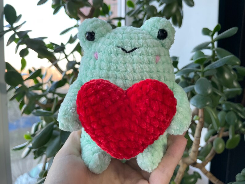 May include: A green crocheted frog holding a red crocheted heart. The frog has black eyes and a black nose.