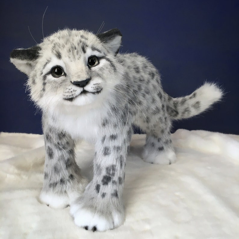 May include: A white and gray plush snow leopard cub with black spots. The cub is standing on a white fluffy surface and looking up at the camera.