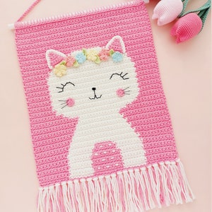 Peut inclure: Une tapisserie murale en crochet rose avec un chat en crochet blanc portant une couronne de fleurs. Le chat a une expression joyeuse et est entour&eacute; de franges roses.
