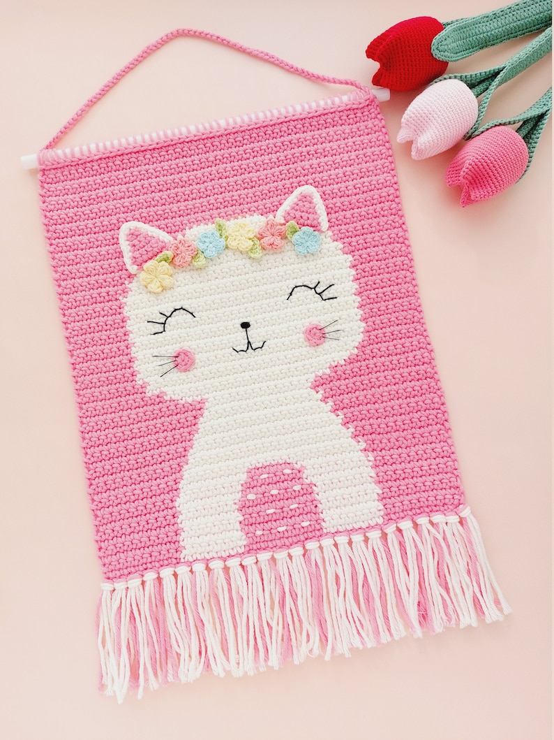 Peut inclure: Une tapisserie murale en crochet rose avec un chat en crochet blanc portant une couronne de fleurs. Le chat a une expression joyeuse et est entour&eacute; de franges roses.