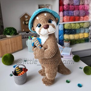 Cute crocheted otter toy holding a colorful striped fish, wearing a soft turquoise bucket hat. Handmade plush animal made with velour yarn – perfect amigurumi decor or gift idea.