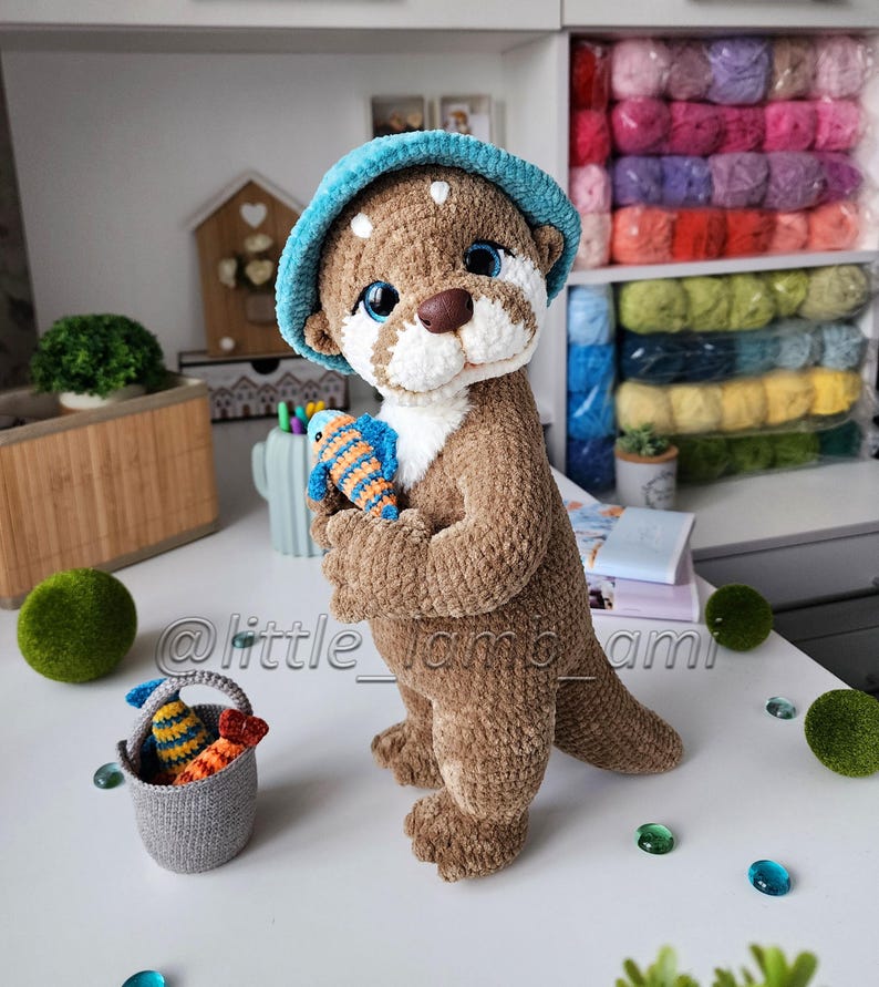 Cute crocheted otter toy holding a colorful striped fish, wearing a soft turquoise bucket hat. Handmade plush animal made with velour yarn – perfect amigurumi decor or gift idea.