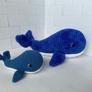 May include: Two crocheted whale plushies, one large and one small, both blue with white accents around the mouth. The whales have black eyes and are made with a textured yarn.