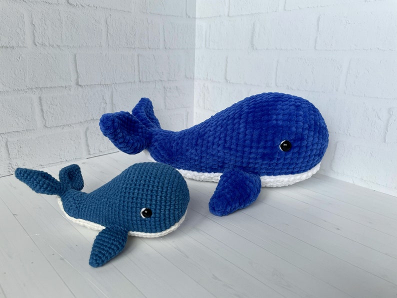 May include: Two crocheted whale plushies, one large and one small, both blue with white accents around the mouth. The whales have black eyes and are made with a textured yarn.