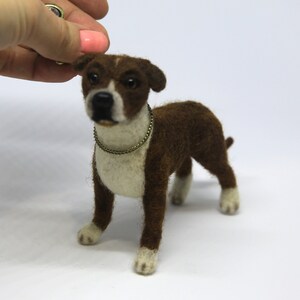 May include: A handcrafted felt dog figurine in brown and white, with a gold-colored collar. The dog has white paws and a white chest. The figurine is standing on a white surface.