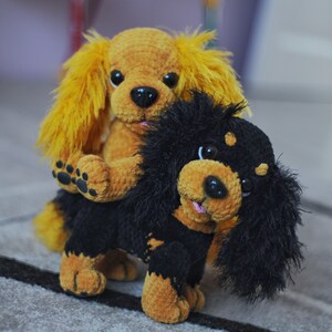 K&ouml;nnte beinhalten: Zwei geh&auml;kelte Stofftiere, ein braunes mit gelbem Fell und ein schwarzes mit braunem Fell. Der braune Hund sitzt auf dem schwarzen Hund.