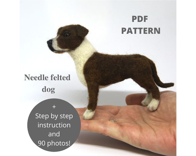 A realistic needle felted Staffordshire-type puppy stands on an open hand against a soft white background. The small handmade dog has brown and white fur, tiny black eyes, and a delicate tail, showing the scale of the finished felted animal. The image also includes the text “PDF PATTERN”, “Needle felted dog”, and “Step by step instruction and 90 photos!”.