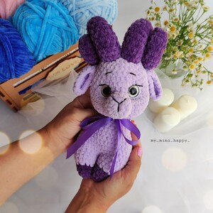 May include: A crocheted purple plush toy with horns, large eyes, and a purple ribbon around its neck. The toy is being held by a person's hands.