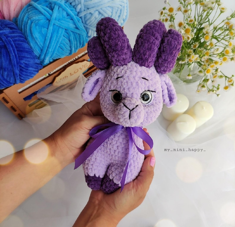 May include: A crocheted purple plush toy with horns, large eyes, and a purple ribbon around its neck. The toy is being held by a person's hands.