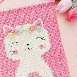 Peut inclure: Une tapisserie murale en crochet rose avec un chat en crochet blanc portant une couronne de fleurs. Le chat a un nez rose et des moustaches noires. La tapisserie murale a une frange blanche et rose en bas.