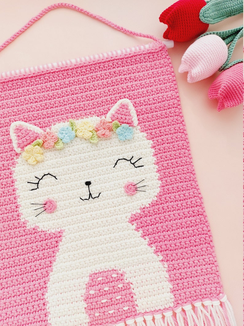 Peut inclure: Une tapisserie murale en crochet rose avec un chat en crochet blanc portant une couronne de fleurs. Le chat a un nez rose et des moustaches noires. La tapisserie murale a une frange blanche et rose en bas.
