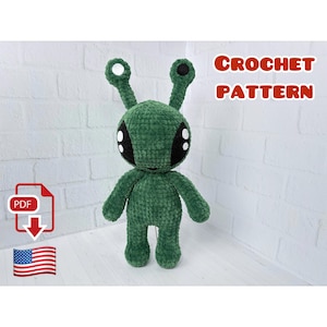 Op de afbeelding: Een groene gehaakte alienpop met grote zwarte en witte ogen, twee antennes en kleine armen en benen. De tekst "CROCHET PATTERN" wordt in het rood weergegeven. Een PDF-pictogram en een Amerikaanse vlag zijn ook zichtbaar.