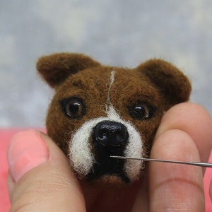 May include: A close-up of a brown and white felted dog head with a black nose and dark eyes. A needle is being used to work on the felted sculpture. The dog has a white stripe down the center of its face.