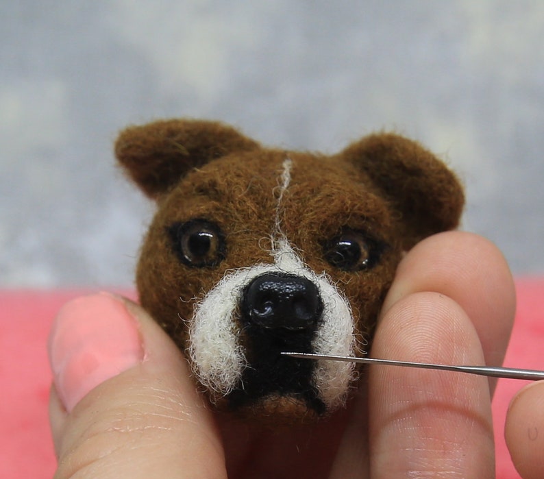 May include: A close-up of a brown and white felted dog head with a black nose and dark eyes. A needle is being used to work on the felted sculpture. The dog has a white stripe down the center of its face.