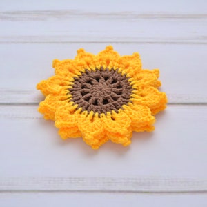 May include: A crocheted sunflower coaster in yellow and brown. The coaster is made of yarn and has a detailed center.