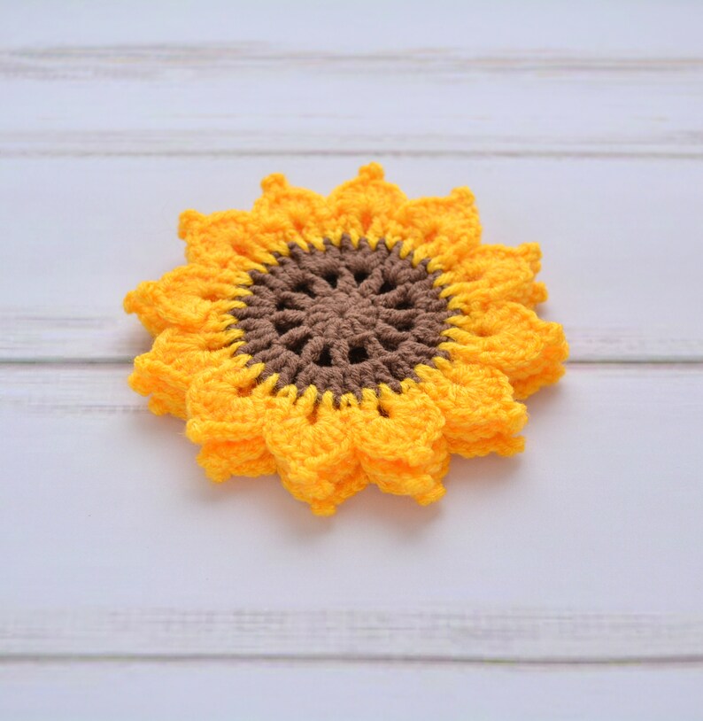 May include: A crocheted sunflower coaster in yellow and brown. The coaster is made of yarn and has a detailed center.
