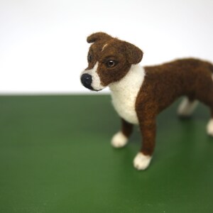 May include: A handcrafted felt dog figurine in brown and white, standing on a green surface. The dog has a white chest and paws, with brown fur on its back and head. The dog's eyes are visible, and it has a black nose.