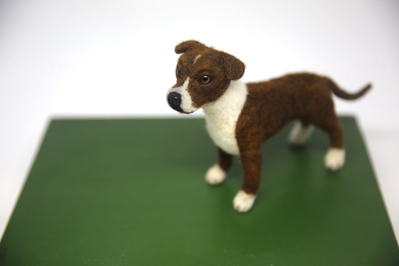 May include: A handcrafted felt dog figurine in brown and white, standing on a green surface. The dog has a white chest and paws, with brown fur on its back and head. The dog's eyes are visible, and it has a black nose.