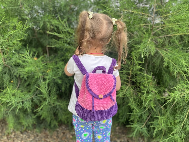 Pu&ograve; includere: Una bambina indossa uno zaino in crochet rosa e viola. Lo zaino ha una chiusura a bottone e una piccola tasca sul davanti.