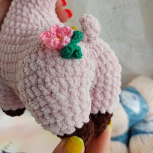 May include: A pink crocheted stuffed animal with brown feet and a flower on its back. The flower is pink with green leaves.