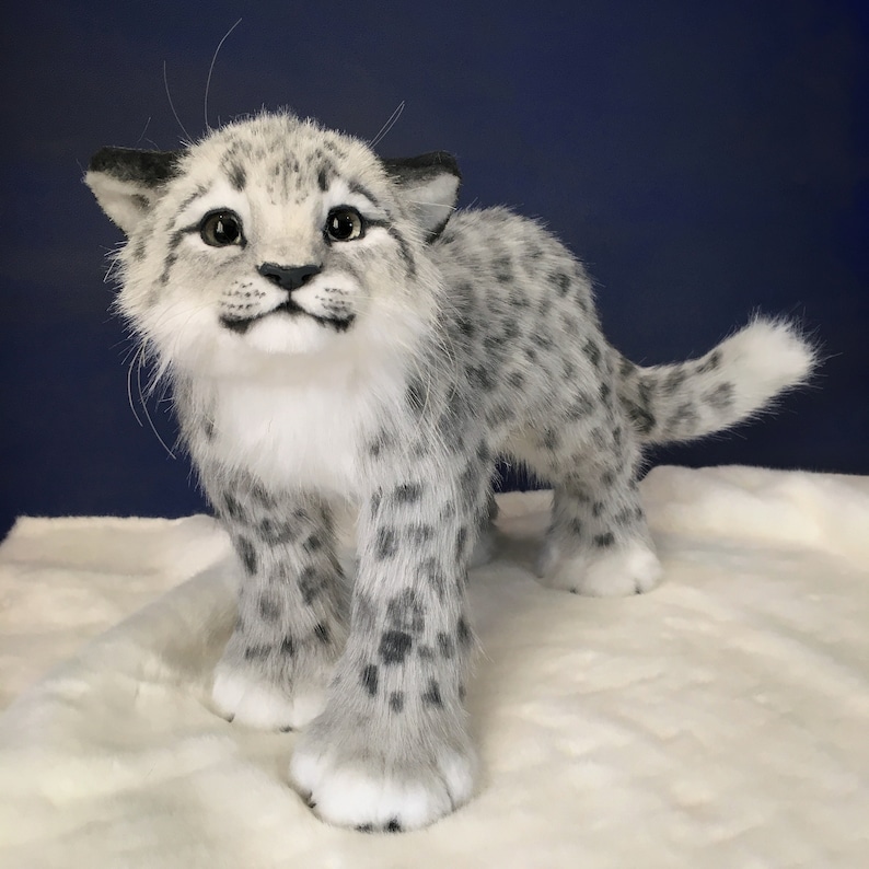 May include: A white and gray plush snow leopard cub with black spots. The cub is standing on a white fluffy surface and looking up at the camera with a playful expression.