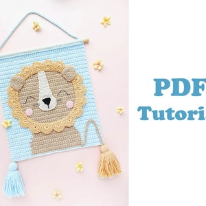 Könnte beinhalten: Gehäkeltes Löwen-Wandbehang mit hellblauem Hintergrund und einem Holzstab. Der Löwe hat eine hellbraune Mähne, eine weiße Nase und rosa Wangen. Der Text "PDF Tutorial" befindet sich in der oberen rechten Ecke. Eine hellblaue Quaste und eine hellbraune Quaste hängen unten.