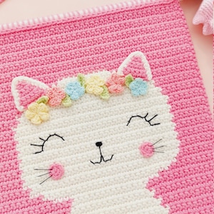 Peut inclure: Une tapisserie murale en crochet rose avec un visage de chat en crochet blanc. Le chat porte une couronne de fleurs avec des fleurs de couleur pastel.