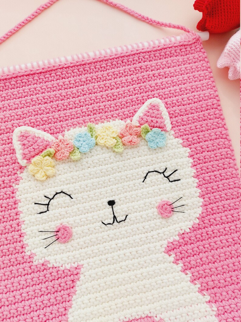 Peut inclure: Une tapisserie murale en crochet rose avec un visage de chat en crochet blanc. Le chat porte une couronne de fleurs avec des fleurs de couleur pastel.