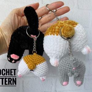以下が含まれることがあります： 3つの編みぐるみ猫のキーホルダー。1つは黒、1つは茶色のしっぽのある白、もう1つはグレー。各キーホルダーにはピンクの肉球が付いており、シルバーのチェーンで繋がっています。