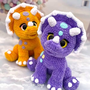 May include: Two crocheted plush triceratops dinosaurs. One is orange with purple spots and the other is purple with blue and yellow spots. Both have white crocheted accents.