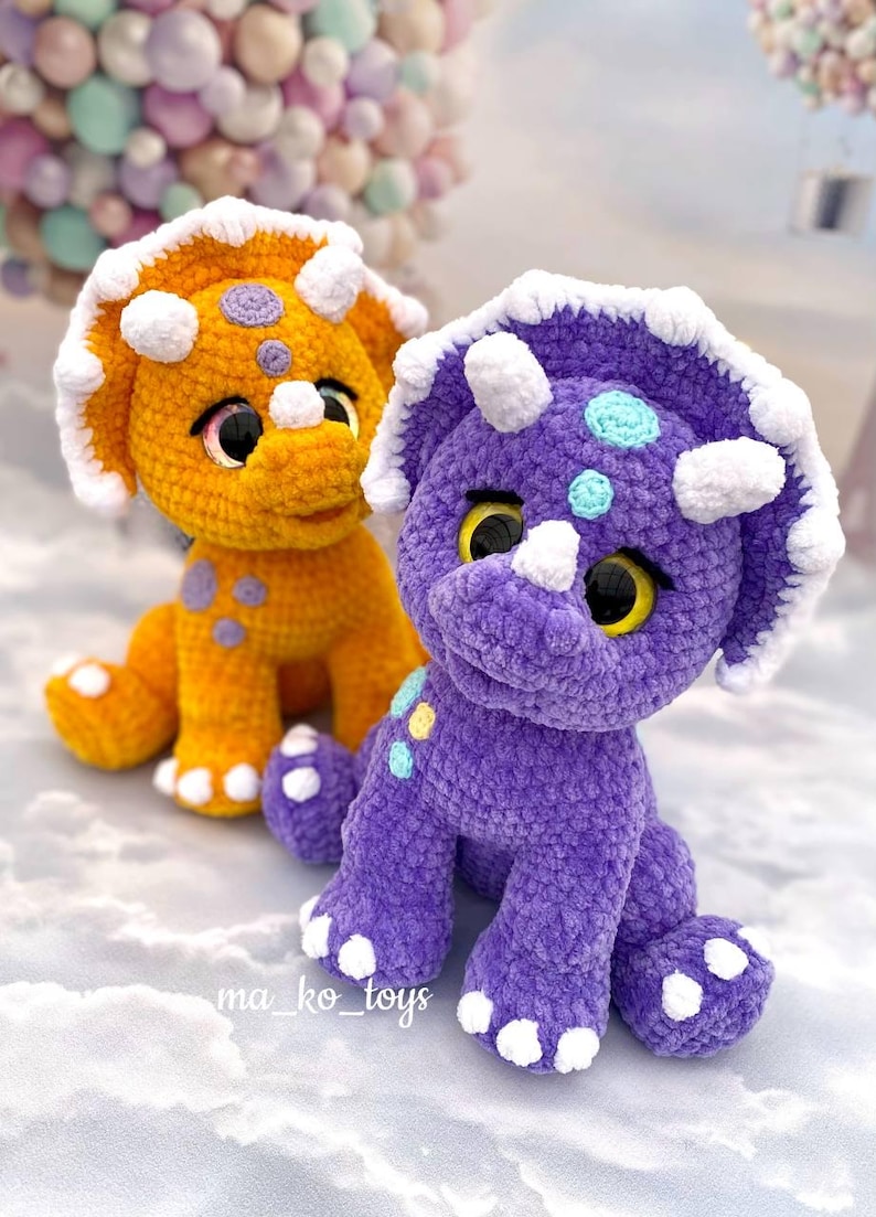 May include: Two crocheted plush triceratops dinosaurs. One is orange with purple spots and the other is purple with blue and yellow spots. Both have white crocheted accents.