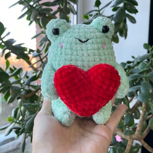May include: A green crocheted frog holding a red crocheted heart. The frog has black eyes and a pink nose.
