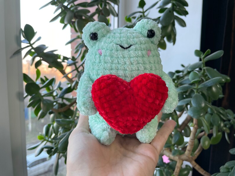 May include: A green crocheted frog holding a red crocheted heart. The frog has black eyes and a pink nose.
