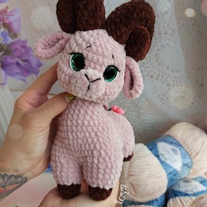 May include: A crocheted pink and brown plush toy ram with green eyes. The toy is being held in a person's hand. The background includes yarn in pink and blue.