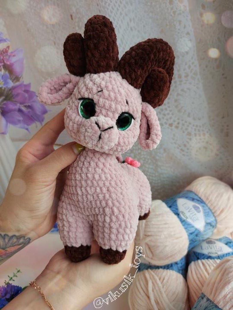 May include: A crocheted pink and brown plush toy ram with green eyes. The toy is being held in a person's hand. The background includes yarn in pink and blue.