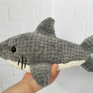Puede incluir: Un juguete de peluche de tibur&oacute;n de crochet gris y blanco. El tibur&oacute;n tiene la barriga blanca y los ojos negros.