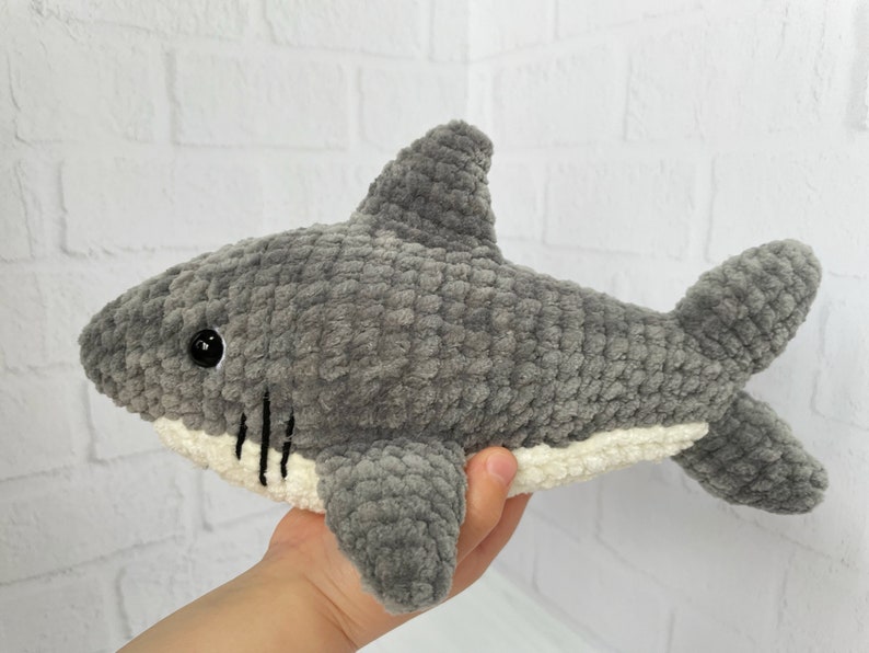 Puede incluir: Un juguete de peluche de tibur&oacute;n de crochet gris y blanco. El tibur&oacute;n tiene la barriga blanca y los ojos negros.