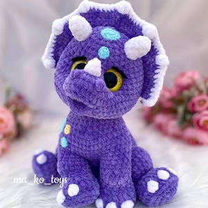 May include: Purple crocheted triceratops dinosaur toy with white horns and feet. The dinosaur has yellow and blue eyes and is decorated with colorful dots.