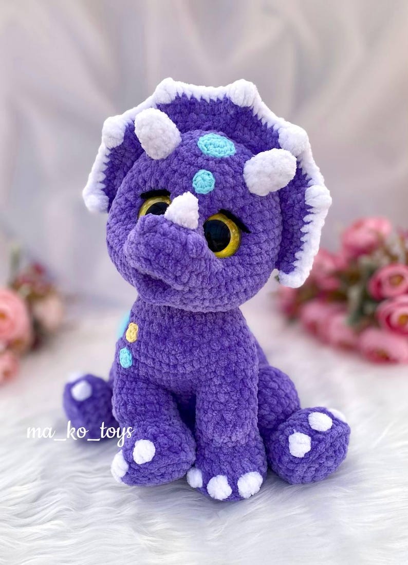 May include: Purple crocheted triceratops dinosaur toy with white horns and feet. The dinosaur has yellow and blue eyes and is decorated with colorful dots.