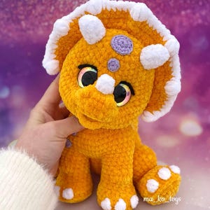May include: A crocheted orange triceratops dinosaur toy with white accents and purple spots. The toy has large, sparkly eyes and a friendly expression.