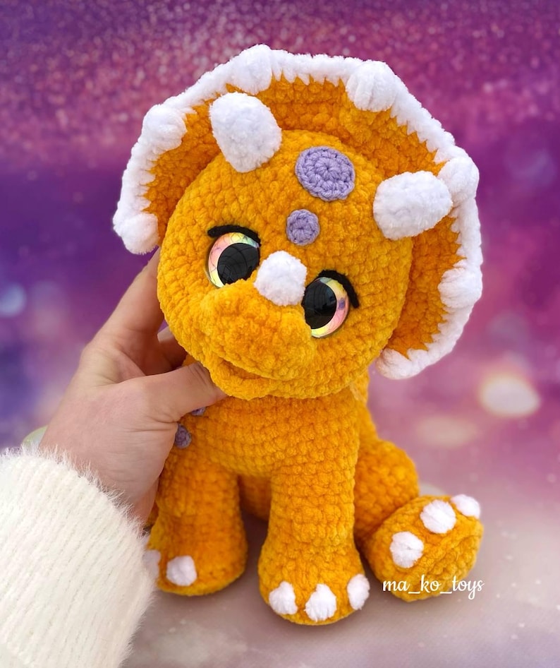 May include: A crocheted orange triceratops dinosaur toy with white accents and purple spots. The toy has large, sparkly eyes and a friendly expression.