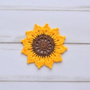 May include: A crocheted sunflower coaster in yellow and brown. The coaster is made of yarn and has a circular shape.